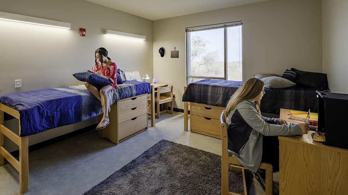 Square dorm room with one individual studying at desk and one studying on bed